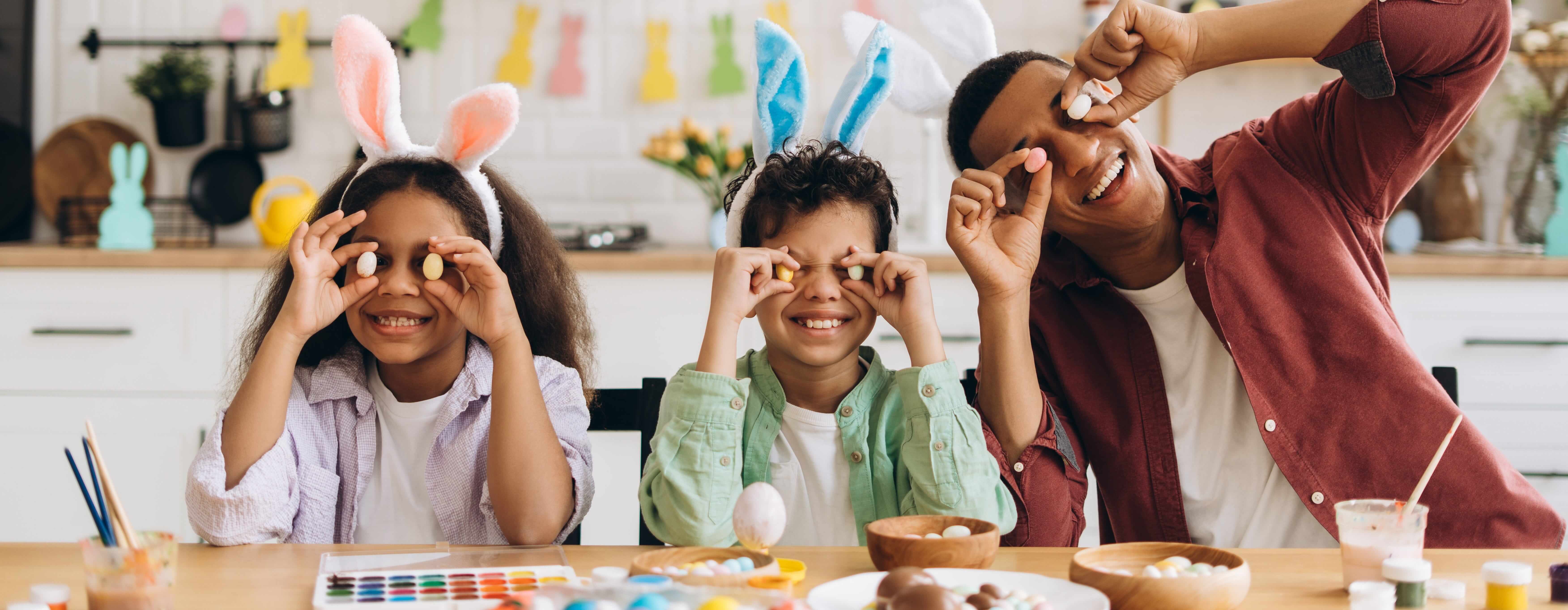 A father on the right hand side with his two children to the left of him, all three sit at a table with the two children wearing bunny ears. all of them are holding up mini eggs to their eyes for the photo.